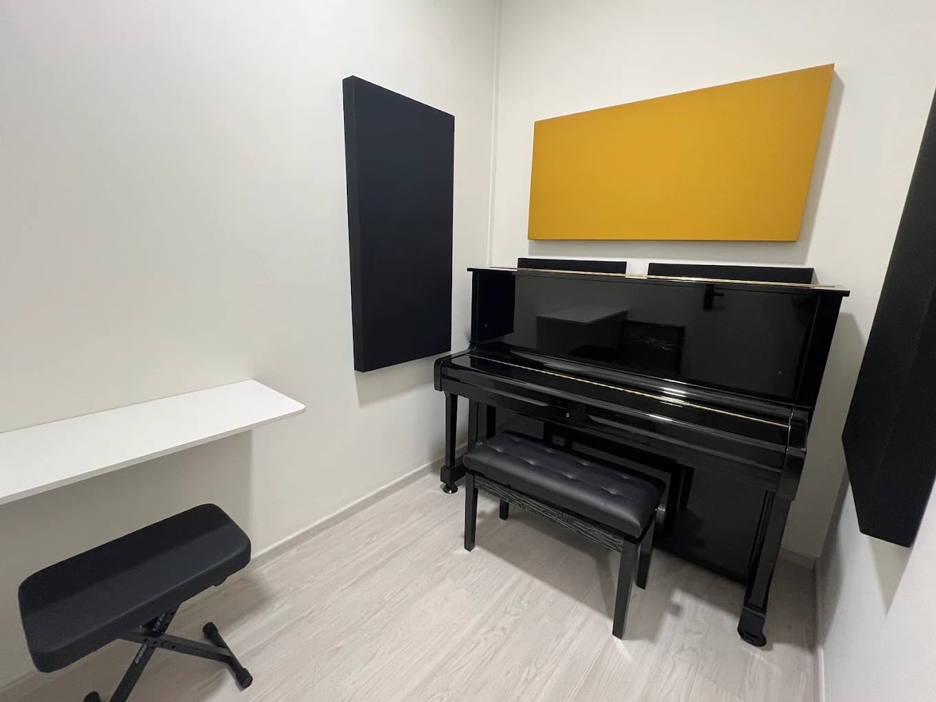 Small individual practice room with a black upright piano, piano bench and a narrow desk, with an acoustic panel on the wall